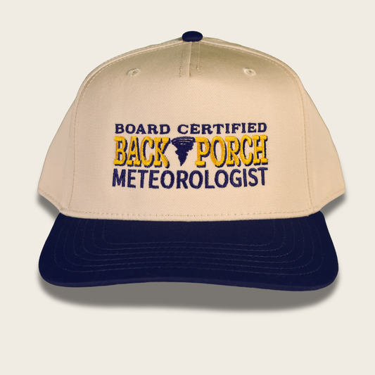 A beige and navy blue baseball cap with embroidered text reading 'BOARD CERTIFIED BACK PORCH METEOROLOGIST' in navy and yellow, with a small tornado icon on the front.