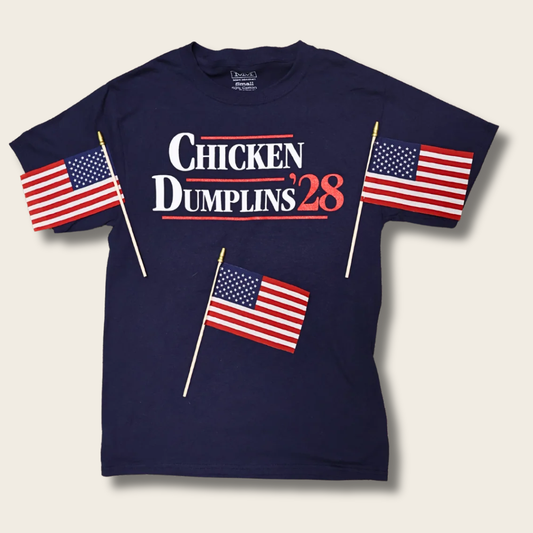A navy blue graphic t-shirt featuring the text 'CHICKEN DUMPLINS '28' in white and red, decorated with three small American flags on sticks printed on the front, set against a light beige background.