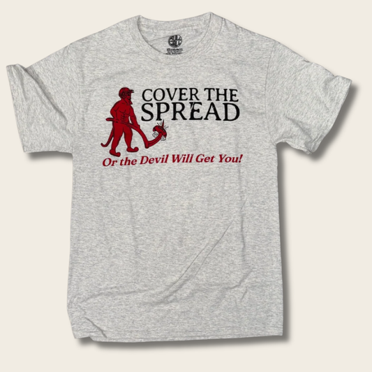 A light heather gray t-shirt with a graphic print featuring a red devil holding a book and the text 'COVER THE SPREAD Or the Devil Will Get You!' in black and red, displayed on a light beige background.