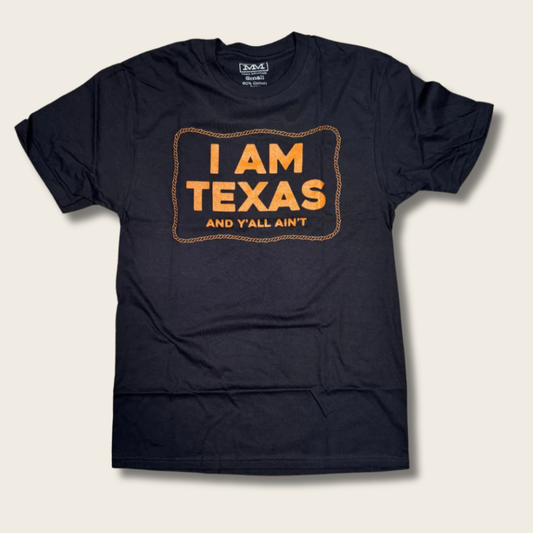 A black short-sleeve t-shirt with a graphic on the chest that reads 'I AM TEXAS AND Y'LL AIN'T' in orange lettering inside a western-style border, displayed flat against a light beige background.