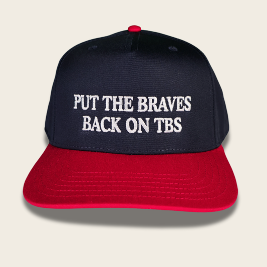 A navy blue and red baseball cap with white embroidered text reading 'PUT THE BRAVES BACK ON TBS' on the front panel, shown against a light beige background.