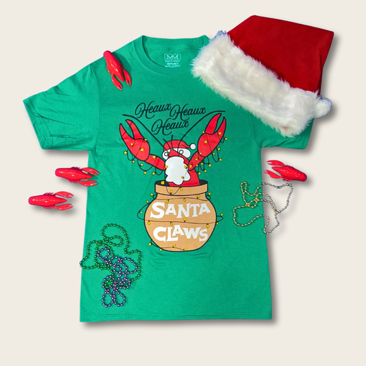 A green short-sleeve T-shirt with a lobster graphic holding a pot labeled 'Santa Claws,' styled with a red Santa hat, red claw-shaped accessories, bead garlands, and holiday lights on a light beige background.