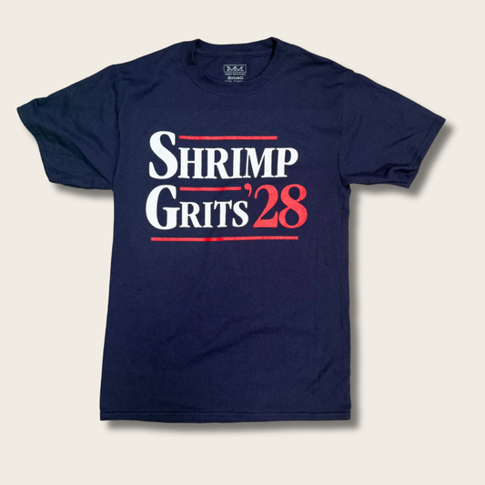 A navy blue short-sleeve t-shirt with a graphic print reading 'SHRIMP GRITS '28' in white and red lettering, displayed flat against a light beige background.