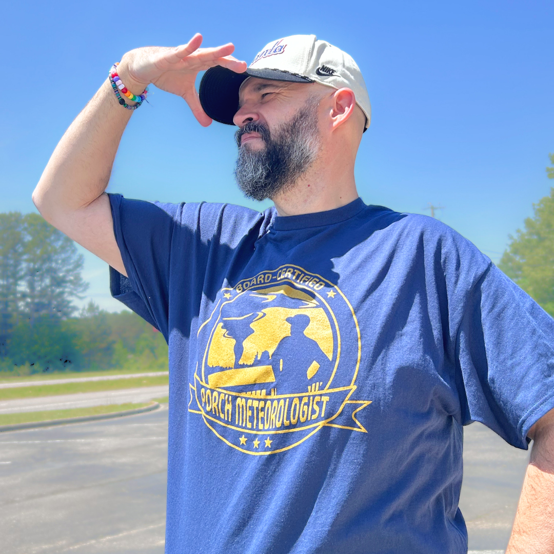 Matt Mitchell wearing a navy blue t-shirt that says "Board-Certified Porch Meteorologist" with graphic of a man watching a tornado, standing outdoors on a sunny day.