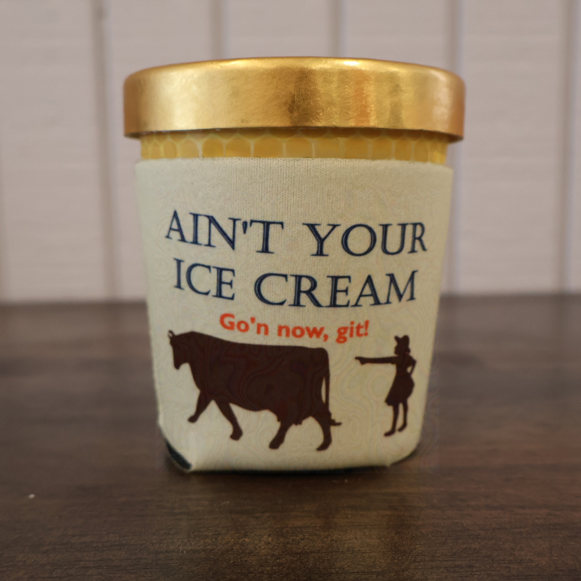 Humorous ice cream koozie with image of girl and cow and text saying "Ain't Your Ice Cream Go'n now, git!" on a wooden surface.