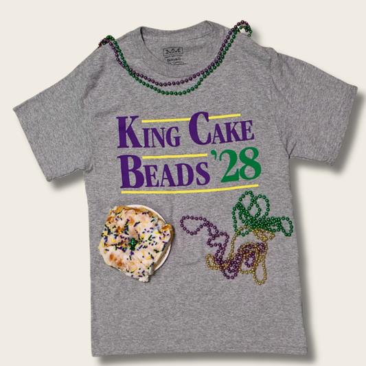 A heather gray short-sleeve t-shirt featuring a 'King Cake Beads '28' graphic with Mardi Gras beads, a donut, and Mardi Gras beads draped around the neckline, set against a light beige background.