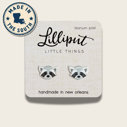 A pair of raccoon stud earrings on a textured display card. The earrings are silver-toned with black and white enamel detailing.