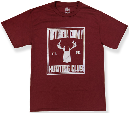 A maroon short-sleeve crewneck t-shirt with a white distressed graphic on the front reading 'Oktibbeha County Stk Mis Hunting Club' around a deer head and cowbell silhouette.